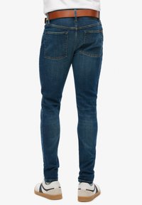 Ciemnoniebieskie dżinsy o wąskim kroju, wyposażone w brązowy skórzany pasek oraz dwie tylne kieszenie, na tle jednolitego białego tła.