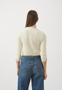 Cremfarbener Strickpullover mit Dreiviertelärmel und gewelltem Ausschnitt, kombiniert mit hochgeschnittenen blauen Jeans aus Denim mit V-förmigen Gesäßtaschen.