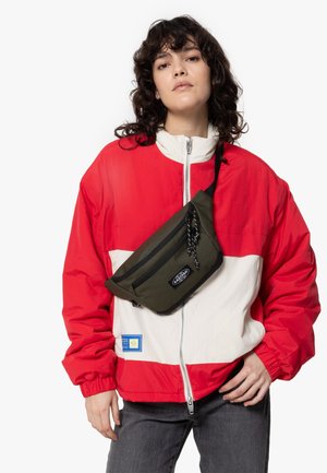 Veste rouge et blanche avec fermeture éclair à l'avant, poignets élastiques et patch. Associée à un sac à taille vert olive avec fermeture éclair et logo.