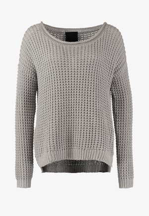 Femme portant un pull en maille gris, un pantalon skinny noir, des bottines noires, et un petit sac bandoulière noir, marchant vers l'avant.