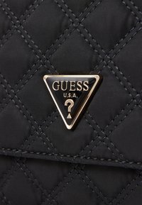 Black quilted fabric with diagonal stitching, featuring a gold triangular Guess logo with a question mark. Smooth texture, modern design.