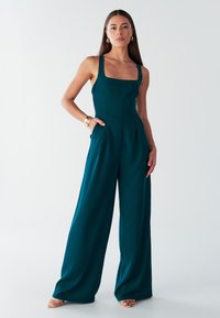 Blågrøn jumpsuit med firkantet udskæring, vide ben og sidelommer. Fremstillet i et glat materiale, med en figursyet overdel og løse bukser.