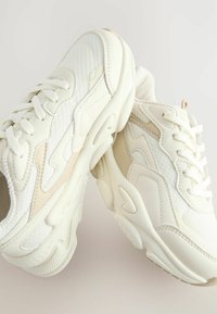 Baskets blanches présentant un mélange de maille texturée et de cuir lisse, avec une semelle épaisse et des accents ton sur ton pour plus de détails.