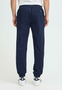Pantalon de jogging bleu marine avec une ceinture élastique, deux poches arrière et des revers côtelés. Porté avec des baskets blanches ornées d'accents noirs.