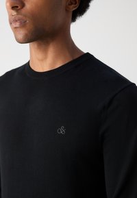 Black cotton crewneck sweater featuring ribbed neckline and a small gray logo on the chest. The fabric appears smooth and lightweight.