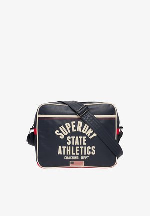 Marineblauwe rechthoekige schoudertas met witte bies, verstelbare riem, tekst "Superdry State Athletics Coaching Dept." en klein Amerikaans vlagembleem.