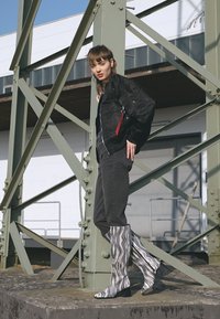Casaco bomber preto, calças cinzas e botas altas com padrões ondulados em preto e branco. Modelo posou contra um fundo industrial.