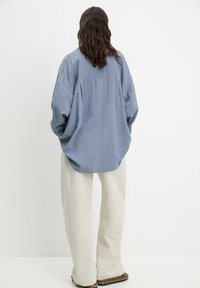 Persona con lunghi capelli mossi che si trova di spalle indossando una camicia blu oversize, pantaloni beige larghi e scarpe marroni con borchie su uno sfondo bianco.