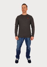 Tricou gri cu mânecă lungă fabricat din bumbac, având un mic logo roșu pe piept, asociat cu blugi albaștri și pantofi sport navy.