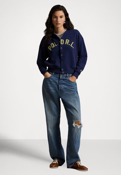 Vrouw draagt een marineblauw gebreid vest met knopen en de tekst "POL O R L", gescheurde brede jeans, bruine veterschoenen, staat met handen in zakken.