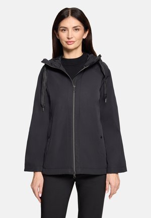 Kobieta ubrana w czarną kurtkę na zamek z kapturem i ściągaczami, stojąca przodem na jednolitym tle.