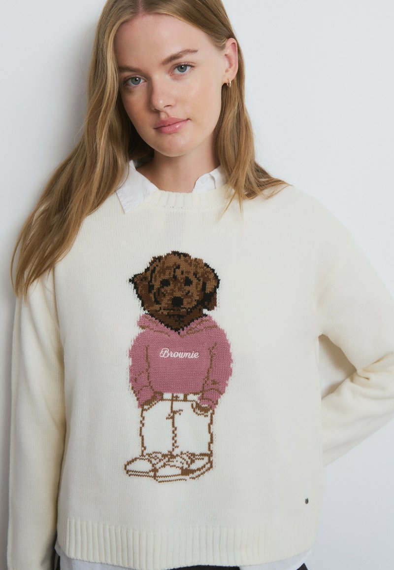 Cremefarbener Pullover mit einem gestrickten Cartoon-Hund, der einen pinkfarbenen Kapuzenpullover mit dem Schriftzug "Brownie" trägt. Der Welpe ist mit weißen Hosen und Turnschuhen dargestellt.