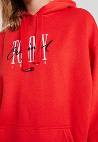 Roter Kapuzenpullover mit weißer und schwarzer bestickter Aufschrift "Tommy Jeans", getragen von einer Person mit hellbraunen Haaren.