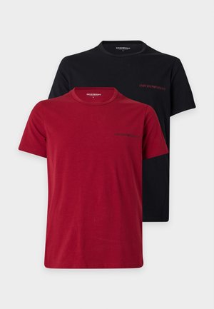 Mann steht barfuß, trägt ein schwarzes T-Shirt mit kurzen Ärmeln und schwarze Hose vor weißem, einfarbigem Hintergrund.