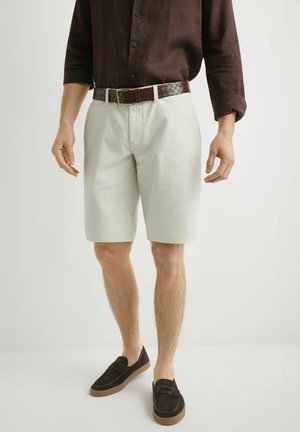 Mężczyzna w kremowych szortach za kolano, ciemnobrązowym plecionym pasku, ciemnobrązowej koszuli z długim rękawem oraz ciemnobrązowych mokasynach z gumowymi podeszwami.