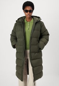 Olive green, knee-length puffer coat with a hood, quilted design, front zipper closure, and side pockets. Worn over a green sweater.
