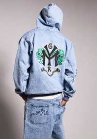 Sudadera de mezclilla con capucha, que presenta un diseño bordado en verde y negro en la parte posterior. La chaqueta tiene un patrón azul tonal y puños acanalados.