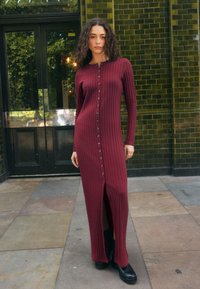 Abito midi color granata a coste con maniche lunghe, bottoni frontali e spacco laterale; indossato con stivaletti neri su una superficie in pietra.