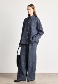 Camisa oversized em mistura de lã texturizada na cor azul-marinho, com grande colarinho e fecho de botões, acompanhada de calças de perna larga. O modelo segura uma carteira preta.