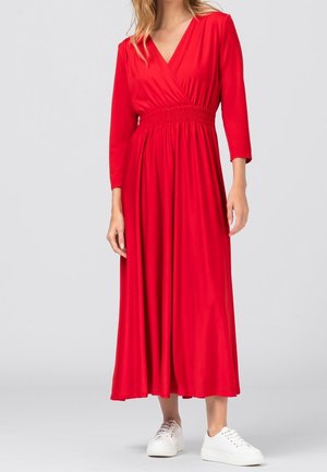 Femme portant une longue robe portefeuille rouge avec des manches trois quarts et des baskets blanches, debout devant un fond clair uni.