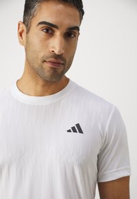 Valge sportlik T-särk tekstuuriga mustriga. Vasakul rinnakorvil on must logo. Lühikesed varrukad ja ümmargune kaelus.