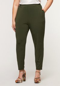 Nainen, jolla on yllään oliivinvihreät slim-fit-housut, joissa on joustava vyötärö ja korkeakorkoiset käärmeennahkakuosiset platform-sandaalit, toinen käsi taskussa.