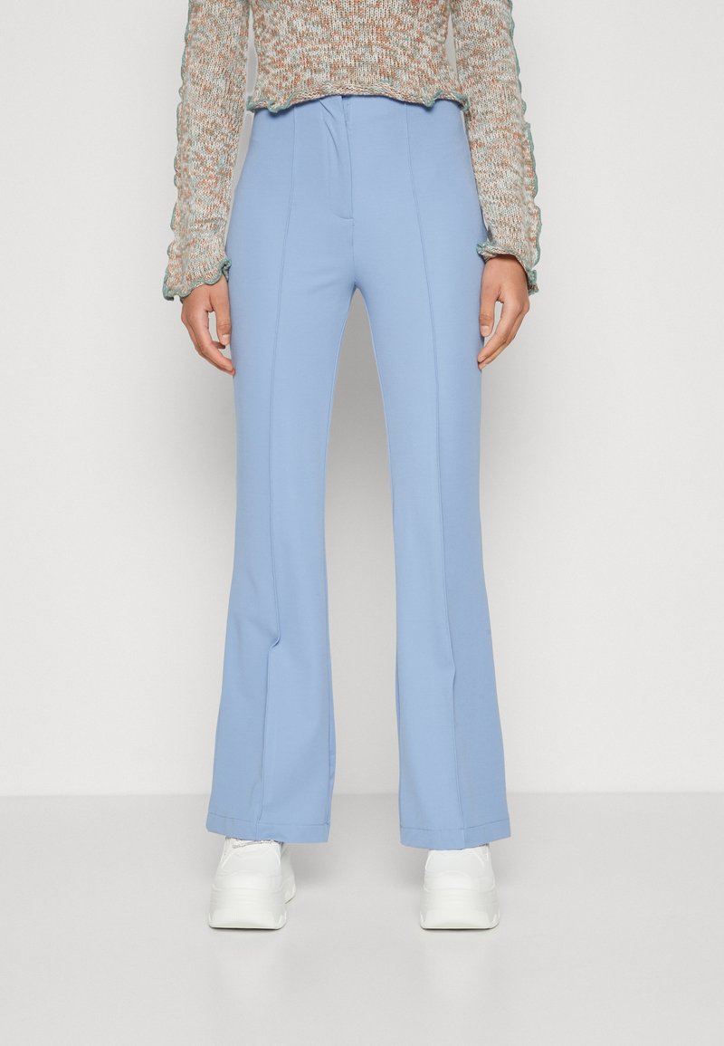 Femme portant un pantalon évasé bleu clair, des baskets à plateforme blanches et un pull en maille multicolore, debout devant un fond uni.