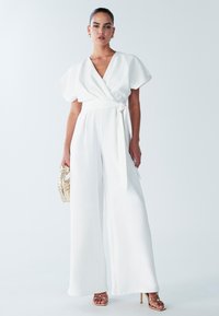 Witte wikkeljumpsuit met korte, wijde mouwen en een ceintuur in de taille. Voorzien van broek met wijde pijpen en een gladde, zijdeachtige textuur. Gecombineerd met een geweven tas.