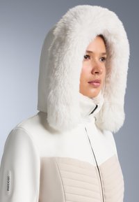 Bijela vanjska jakna s pufastim bijelim umetkom od umjetnog krzna na kapuljači i bež prošivenim panelima. Ima zatvaranje na prednjem dijelu sa patentnim zatvaračem i RECCO logotip na rukavu.