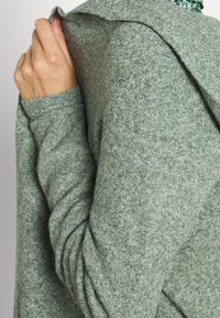 Zelený texturovaný svetr s kapucí vyrobený z měkké pletené látky. Design zahrnuje žebrované manžety a volný střih.