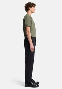Olivegrünes T-Shirt mit kurzen Ärmeln und entspanntem Schnitt, kombiniert mit schwarzen, maßgeschneiderten Hosen und schwarzen Schnürschuhen. Seitenansicht.