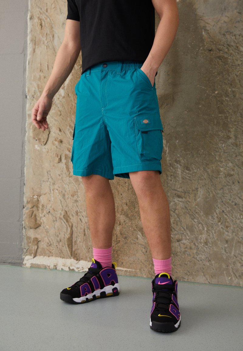 Shorts cargo turquoise avec poches latérales, dotés d'une fermeture à boutons. Portés avec des baskets noires et violettes et des chaussettes roses.
