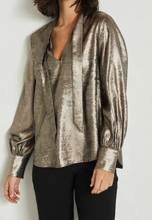 Kvinde iført en metallic bronze langærmet bluse med V-udskæring og bundet tørreldetalje, kombineret med sorte bukser mod en ensfarvet baggrund.