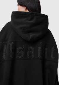 Svart sweatshirt med en stor huva, texturerad sömnad och ordet "Allsaint" i en fet, tonlad stil över ryggen.