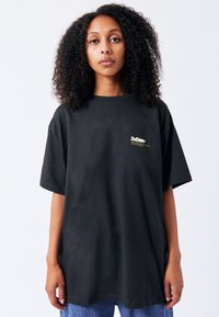 Černé oversized tričko vyrobené z bavlny, s malým zeleným logem na levé části hrudi. Krátké rukávy a uvolněný střih.