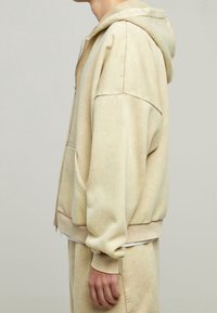 Felpa con zip di colore beige chiaro, realizzata in tessuto morbido, con un cappuccio ampio, tasche frontali e polsini e orlo a coste.