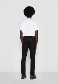Homme avec coiffure afro portant une chemise blanche à manches courtes, un pantalon noir et des chaussures noires, debout face à un fond uni.