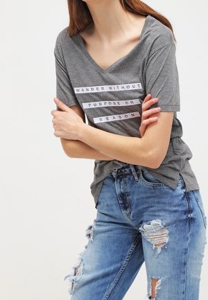 Kvinde iført en grå V-udskåret T-shirt med teksten "VANDR UDEN FORMÅL ELLER GRUND" og slidte blå jeans, armene krydset over brystet.