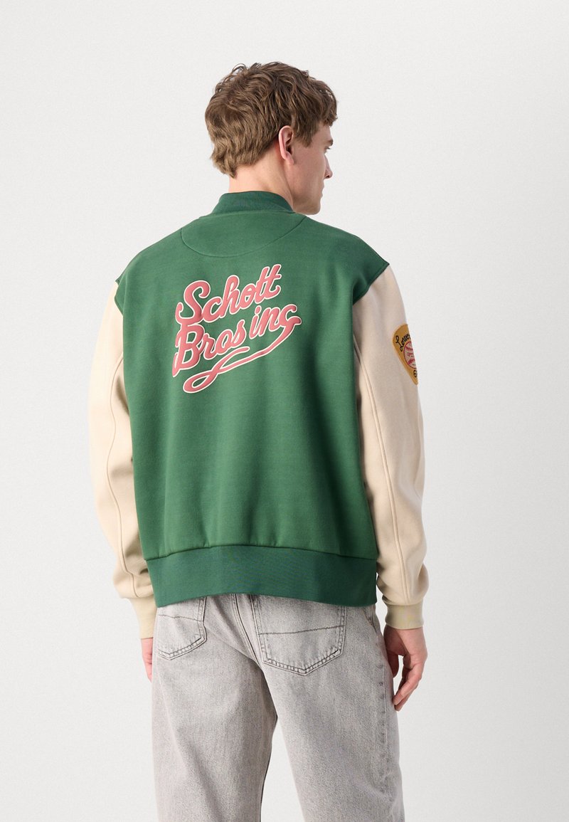 Veste varsity verte et crème avec col et poignets côtelés, affichant "Schott Bros Inc" en cursive rose à l'arrière et un détail de patch sur la manche.