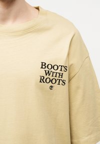 Ανοιχτό μπεζ μπλουζάκι με μαύρο κείμενο «BOOTS WITH ROOTS» και μικρό λογότυπο δέντρου, φορεμένο από ένα άτομο μπροστά σε απλό φόντο.