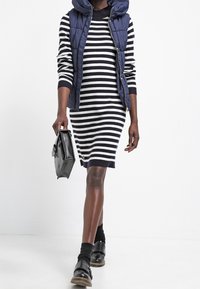 Randig klänning i marinblått och vitt med en figursydd form, kombinerad med en puffig marinblå väst och svarta skor, håller en svart handväska.