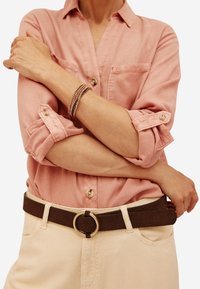 Camicetta rosa chiaro con maniche arrotolate, abbinata a una cintura marrone con una fibbia circolare dorata, indossata con pantaloni chiari.
