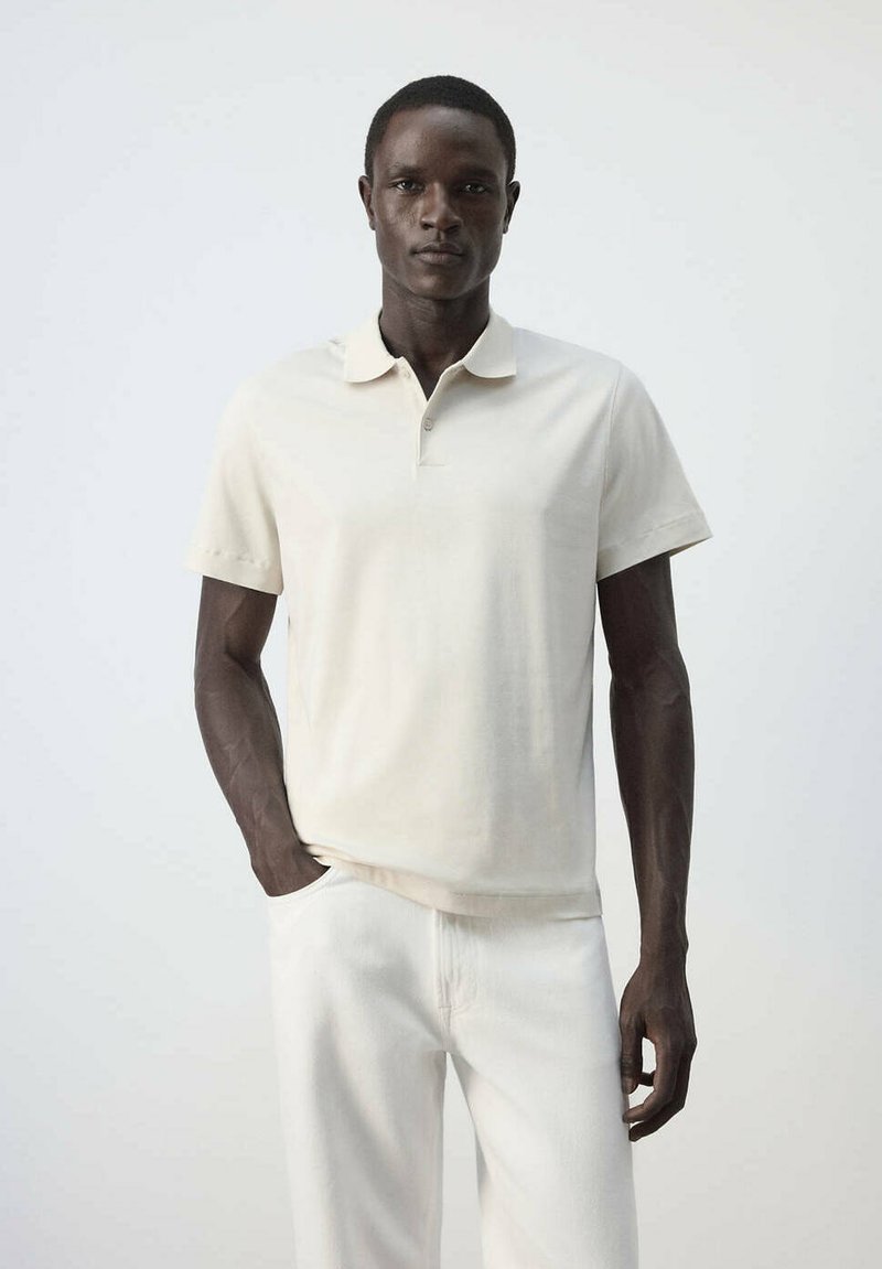 Homme à la peau foncée portant un polo beige à manches courtes et un pantalon blanc, debout devant un fond clair uni.