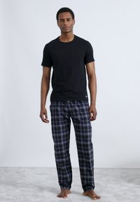 Černé bavlněné tričko ve spojení s tmavě modrými károvanými pyžamovými kalhotami s bílými a fialovými pruhy. Model stojí bosý na světle šedém povrchu.