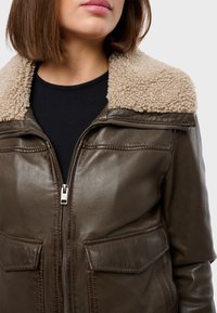 Brun läderjacka med en beige fårskinnskrage, frontdragkedja och två fickor med lappar. Slät textur med kontrasterande sömdetaljer.