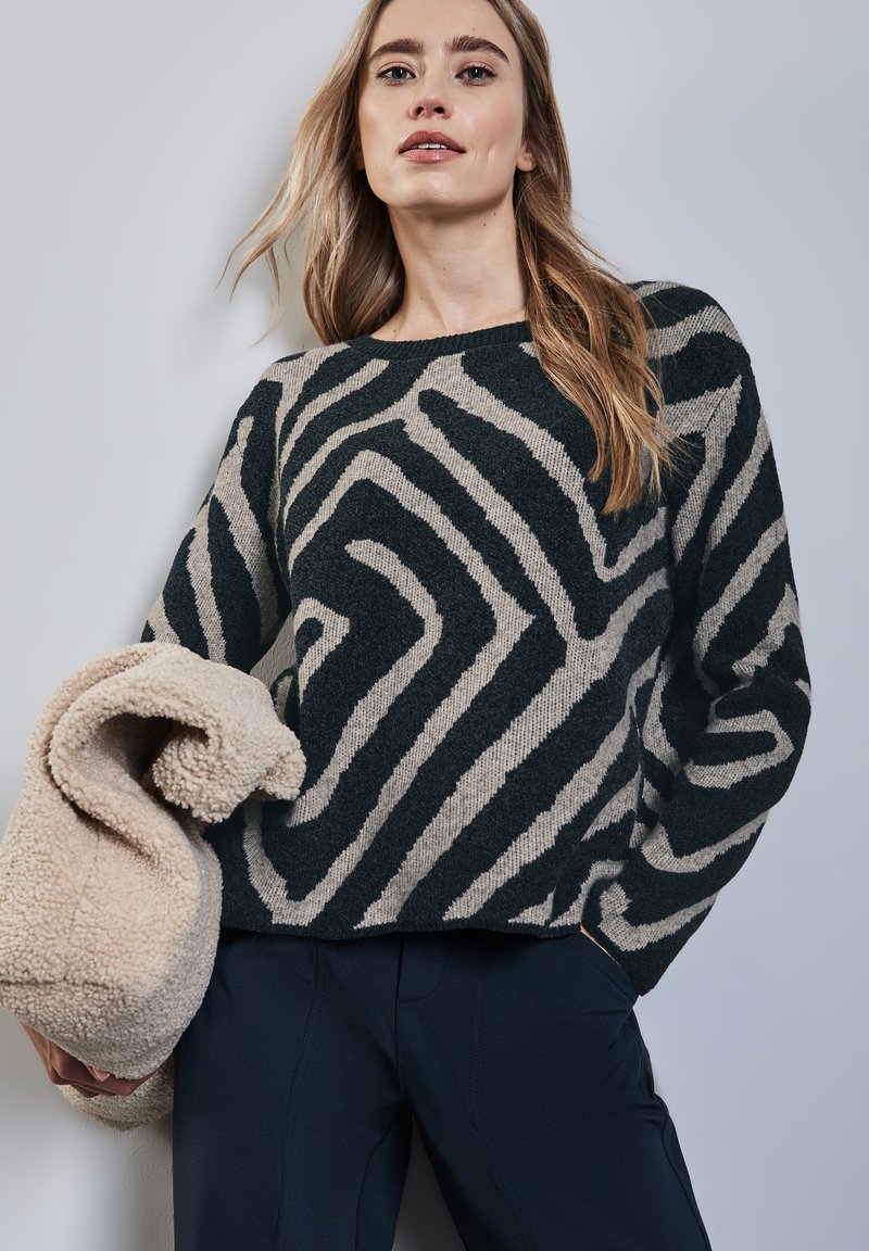 Maglione verde scuro e beige con motivo astratto a zebre, realizzato in tessuto a maglia testurizzato. Modello che tiene un fleece color crema.