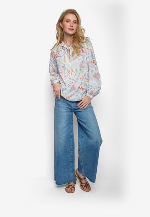 Helle, gemusterte Bluse mit Knoten am Hals, kombiniert mit weit geschnittenen blauen Jeans. Sandalen runden das Outfit ab.