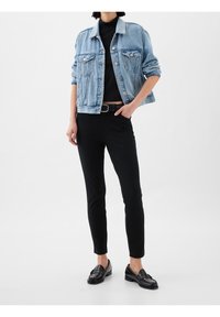 Personne portant une veste en jean bleu clair, un pantalon noir ajusté, une ceinture noire et des mocassins noirs, posant devant un fond blanc uni.