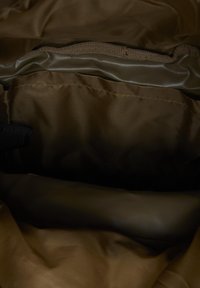 Intérieur d'un sac en tissu brun foncé vide montrant des coutures cousues et une doublure pliée avec une lanière noire partiellement visible à gauche.