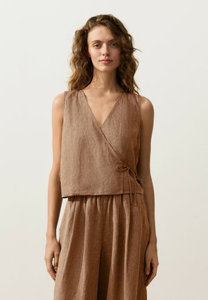 Femeie cu păr creț purtând un top fără mâneci, maro deschis, cu model reiat tip înfășurat și pantaloni largi asortați, pe un fundal simplu, deschis.